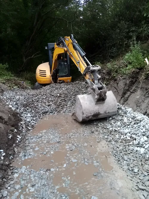 Excavator levelling stone for a new base, in a cutting beside trees.