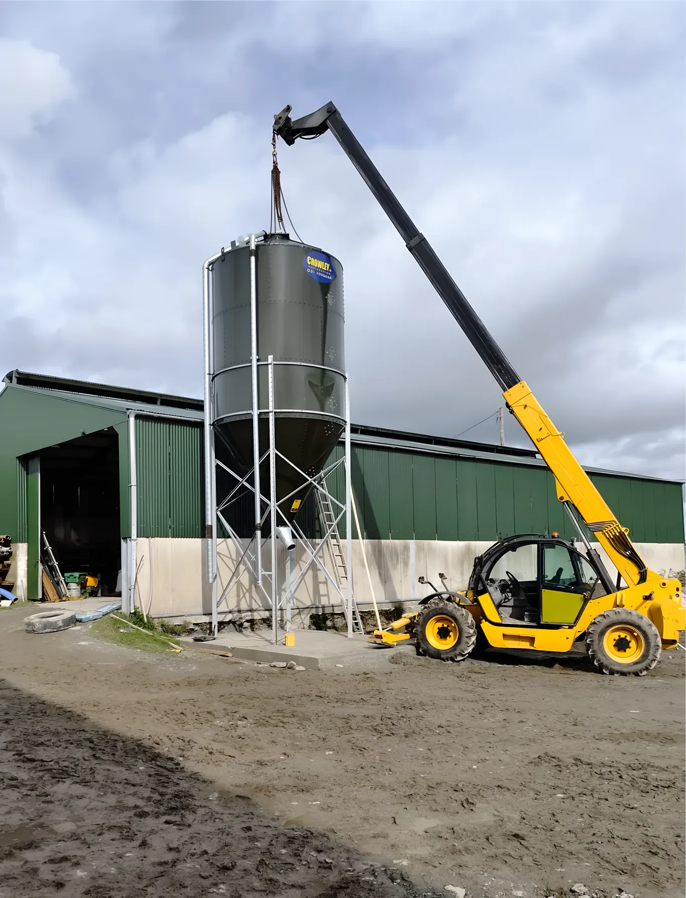 Yellow telescopic loadall lifting a feed silo into place beside a green steel farm shed.